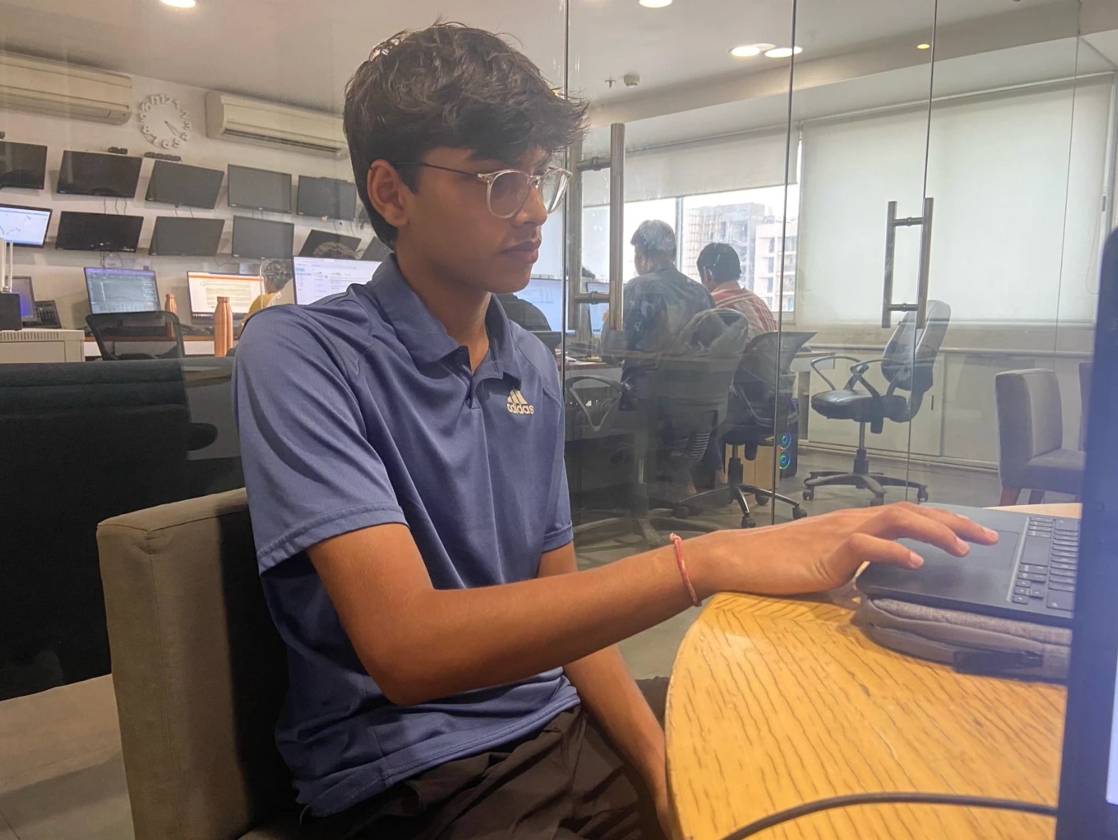 Aarav working on a laptop at the Finesse Stock Broking office, with trading screens on the wall behind him