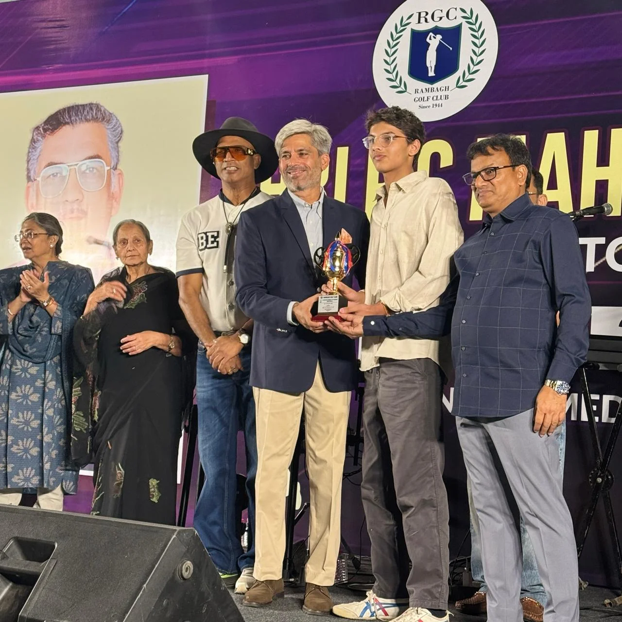 Aarav in a cream shirt receiving a trophy on stage at a Rambagh Golf Club prize-giving