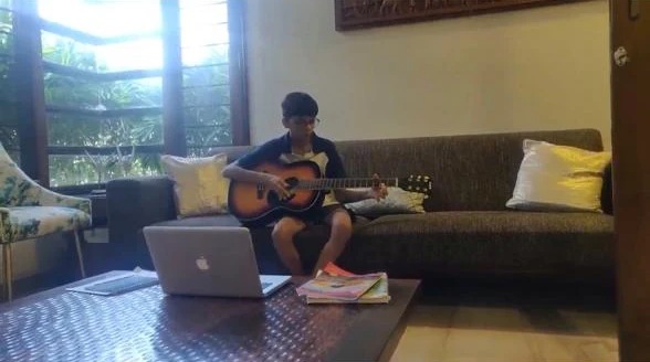 Aarav as a young kid playing an acoustic guitar on a sofa at home, a laptop and songbooks on the table